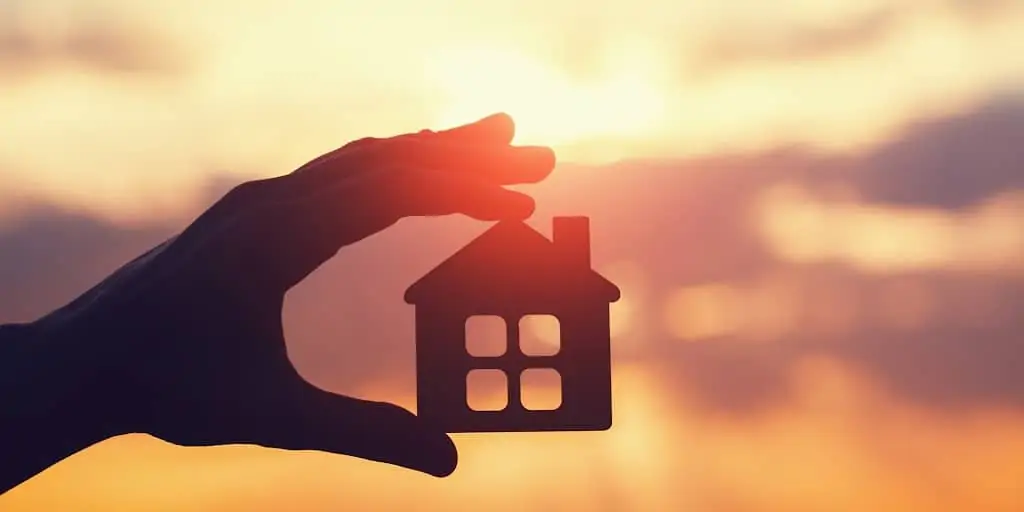 silhouette of hand holding a small wooden house against a sunset