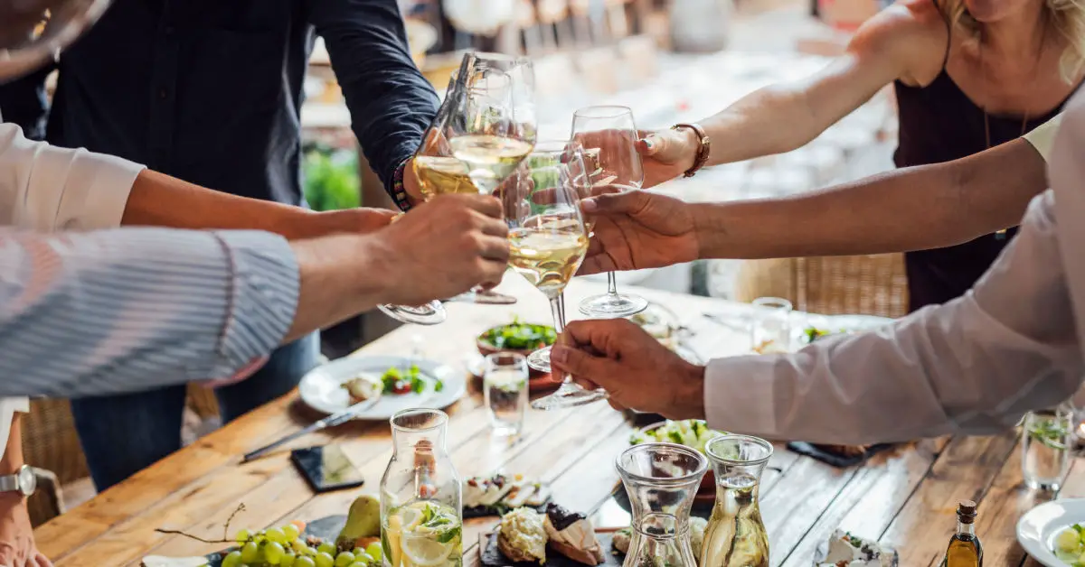 Group of smiling people cheering with wine and rising glasses on celebration at restaurant.